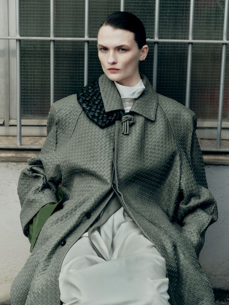 A fashionable adult female is seated wearing a woven grey overcoat with white trousers against an industrial, barred window background.