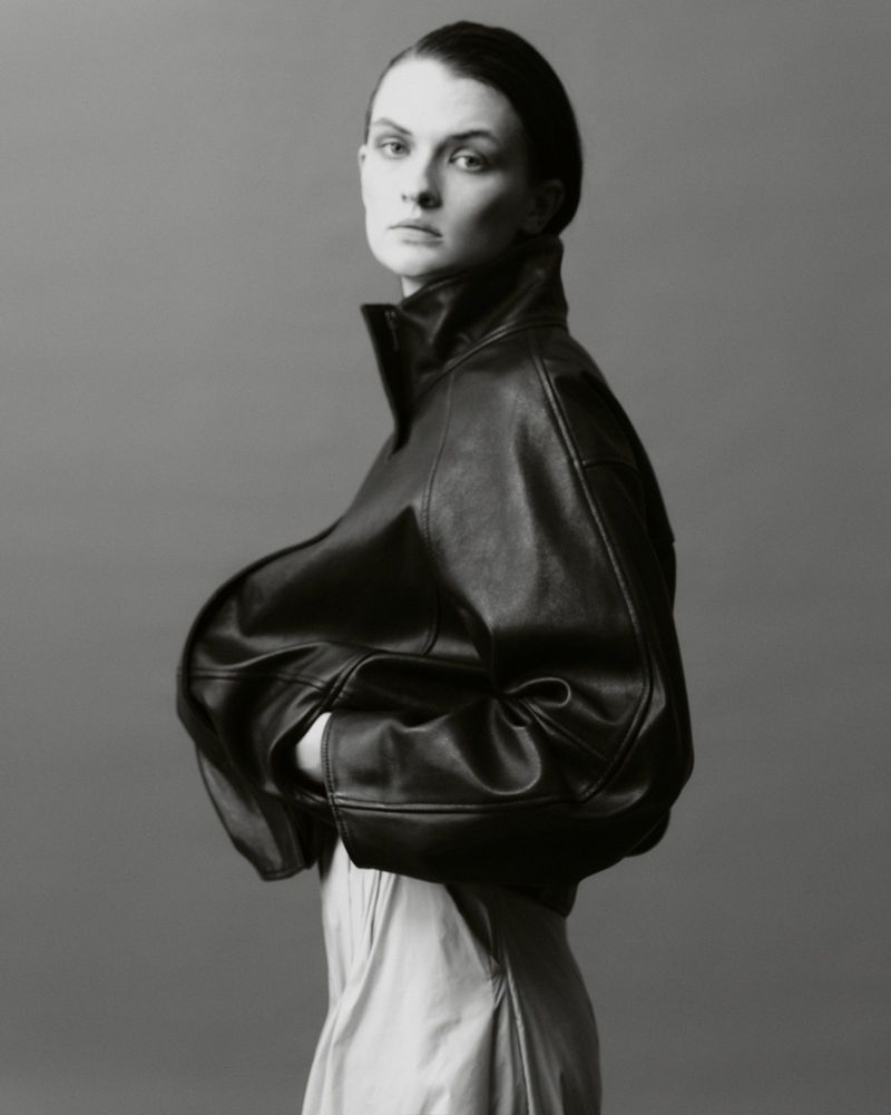 A person with hair pulled back wears an oversized black leather jacket with a high collar, posed against a solid grey background.
