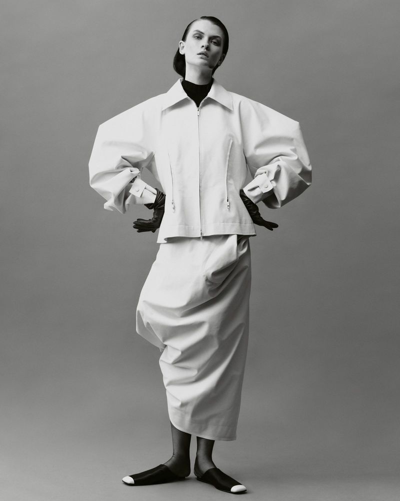 Elegant studio shot of a woman in a white oversized jacket, matching skirt, and black gloves, standing against a grey backdrop.