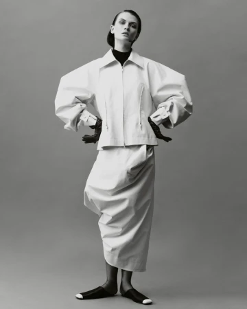 Elegant studio shot of a woman in a white oversized jacket, matching skirt, and black gloves, standing against a grey backdrop.