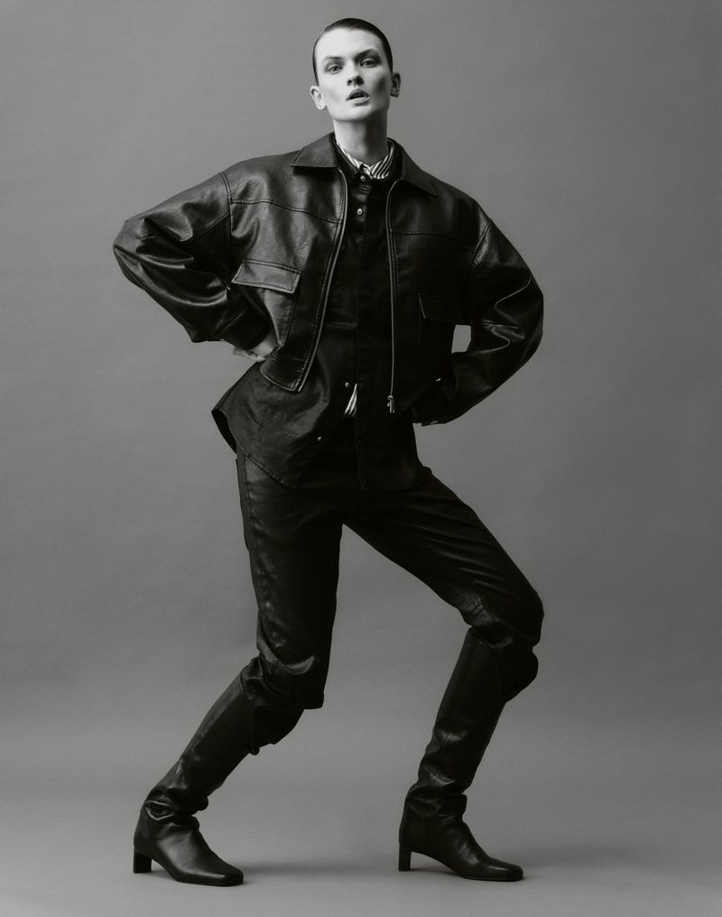 Full-length studio portrait of a female model in a black leather jacket, pants, and heeled boots against a solid gray backdrop.