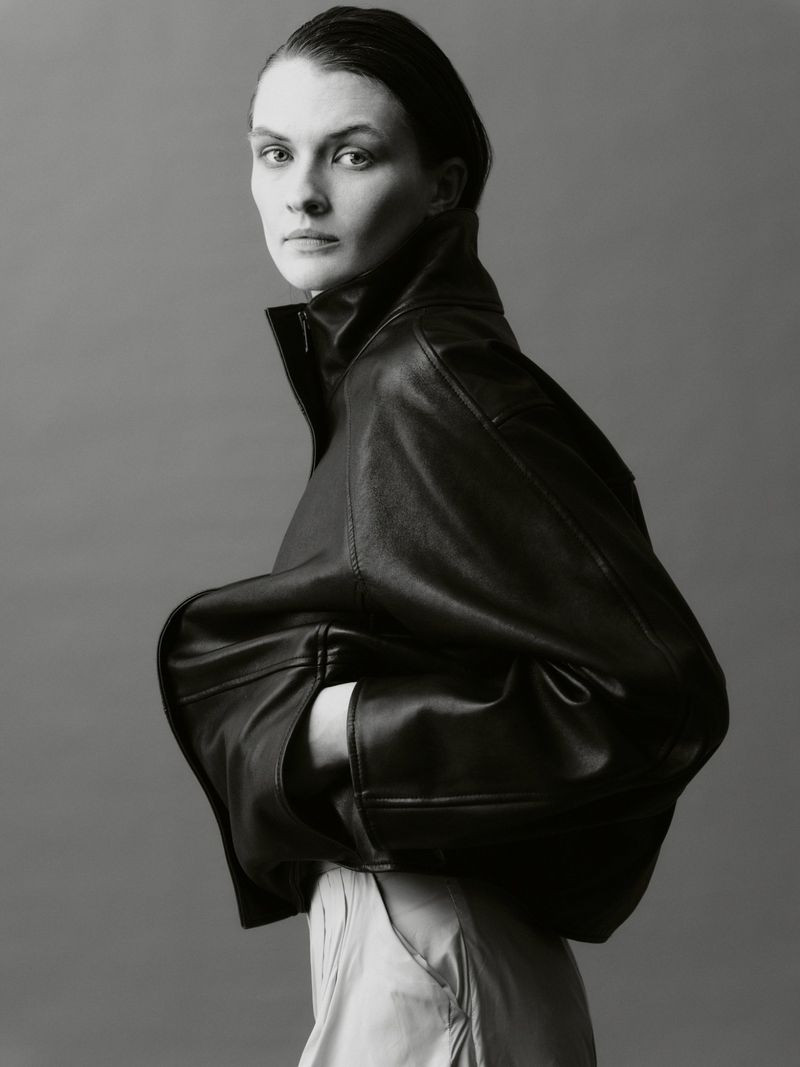 A portrait of a woman wearing an oversized black leather jacket with her hands in the pockets, set against a plain grey background.
