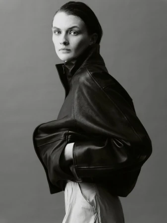 A portrait of a woman wearing an oversized black leather jacket with her hands in the pockets, set against a plain grey background.
