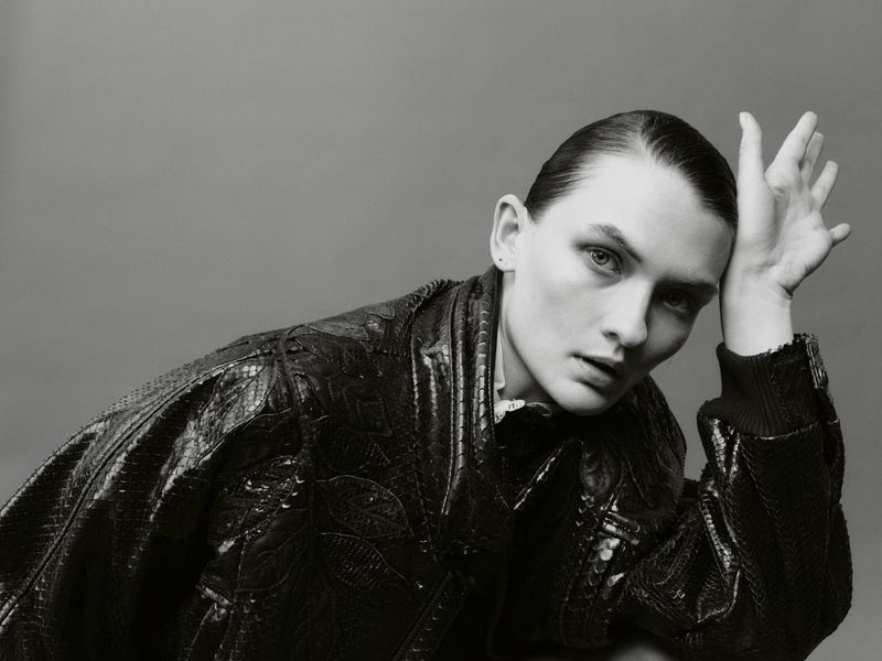 Monochrome portrait of a young adult woman wearing a black textured leather jacket in a studio with gray background.