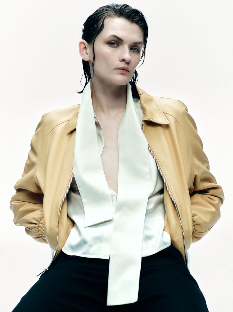 Model with slicked back dark hair wearing a light tan leather jacket over a white blouse with a long scarf against a plain background