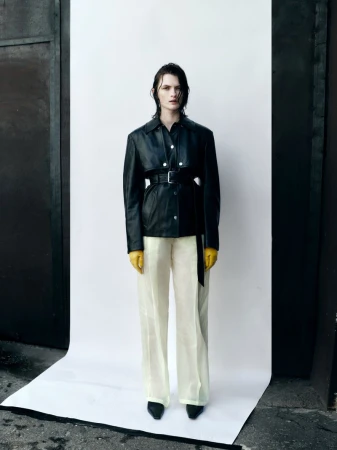 A solo model presents fashion wearing a black jacket, yellow gloves, and light pants against a white backdrop with a dark metal door.