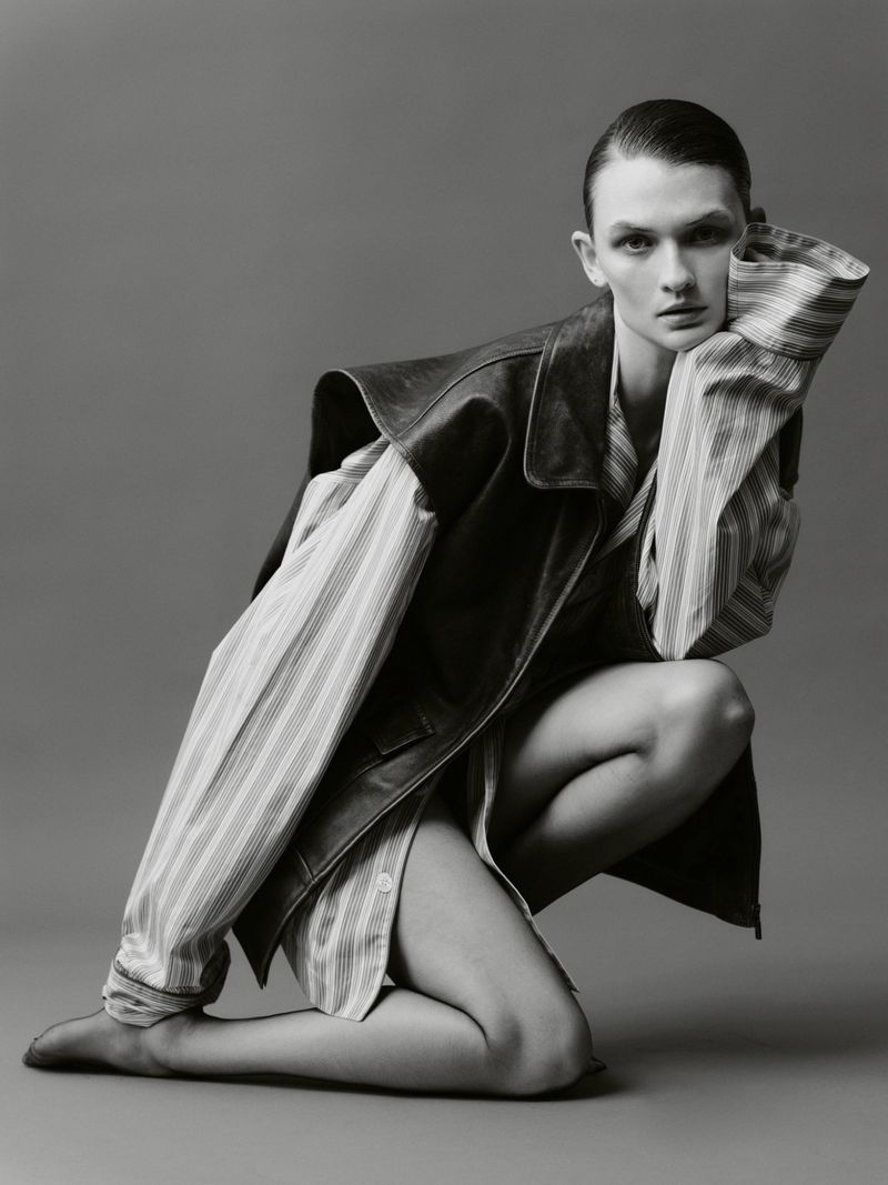 Monochrome studio shot of a woman kneeling in a striped shirt and vest, hand to face, neutral expression against a grey backdrop.