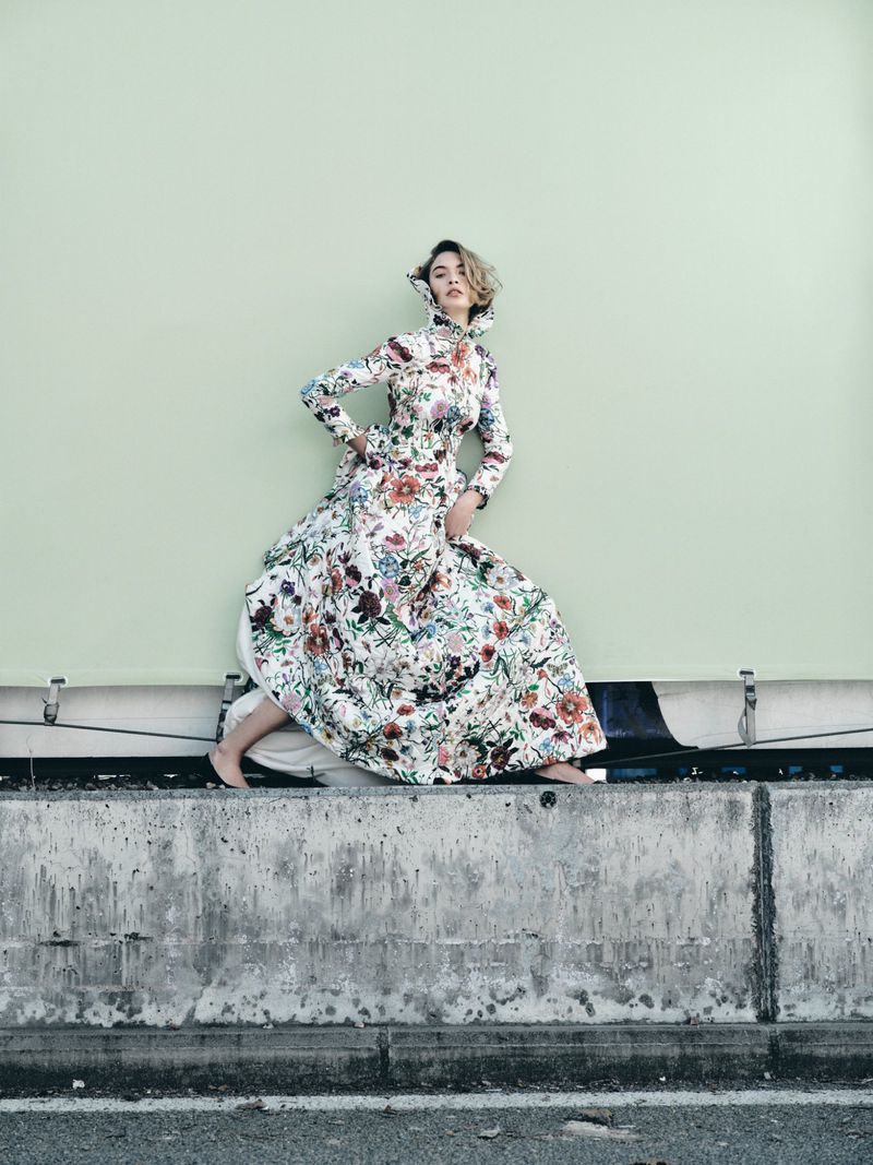 Woman in floral dress poses on concrete barrier against a light green wall, showcasing fashion and outdoor style.