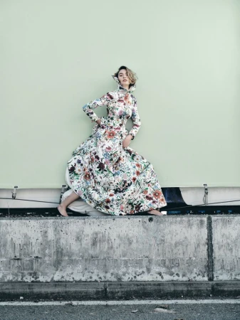 Woman in floral dress poses on concrete barrier against a light green wall, showcasing fashion and outdoor style.