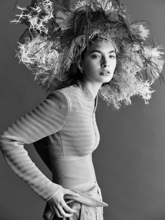 Black and white portrait of a woman with an elaborate feathered headpiece, wearing a textured jacket and skirt, in a neutral setting.