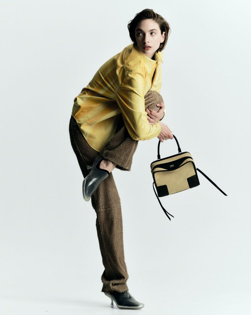 A woman in a yellow shirt and brown pants strikes a pose in a studio with a purse, set against a bright white background.