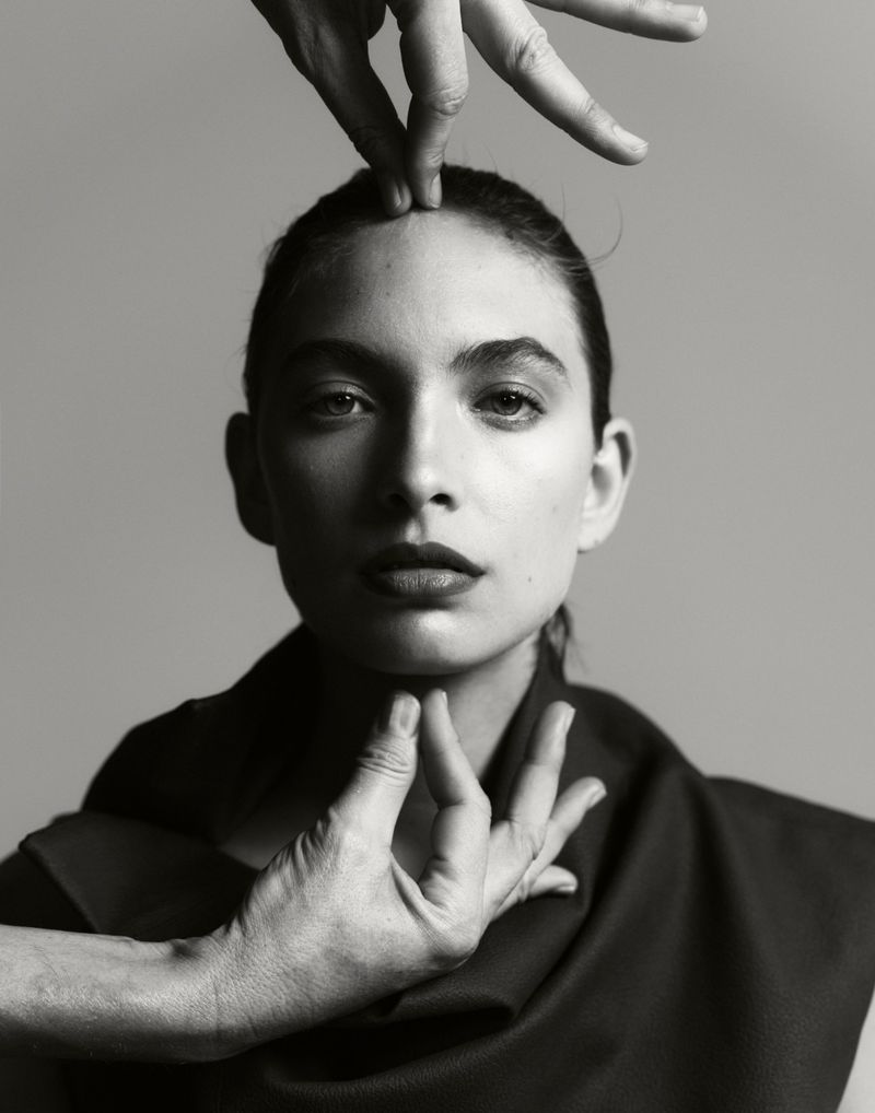 Striking portrait of a woman with hands framing her face against a grey backdrop, creating a dramatic and artistic composition.