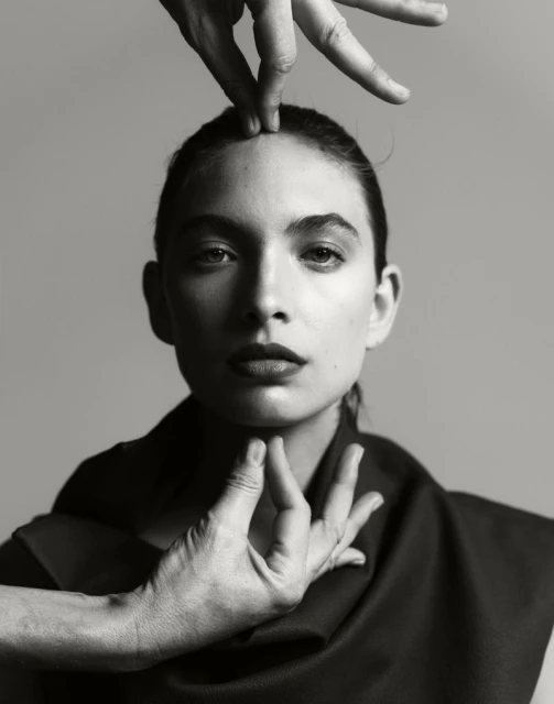 Striking portrait of a woman with hands framing her face against a grey backdrop, creating a dramatic and artistic composition.