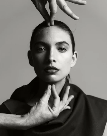 Striking portrait of a woman with hands framing her face against a grey backdrop, creating a dramatic and artistic composition.