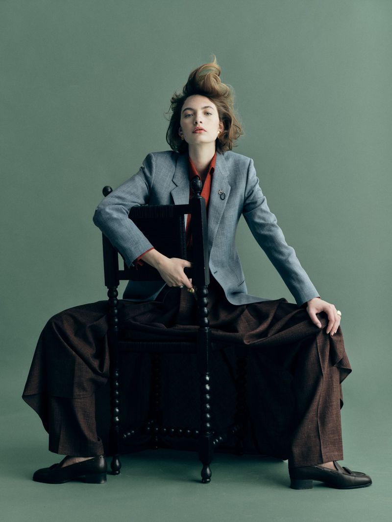 Adult woman sits backward on a black chair in a light blue suit jacket and brown skirt, against a green backdrop in a studio shot.