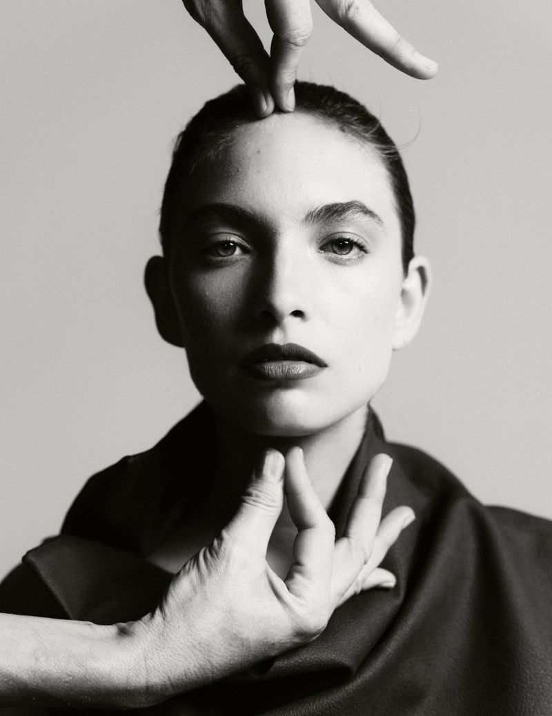 Portrait of a woman with dark hair pulled back, framed by two hands touching her forehead and chin, monochrome shot against grey background.