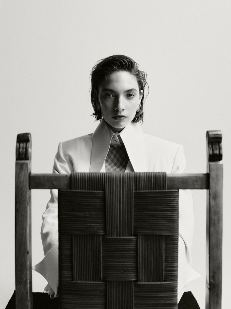 Elegant studio portrait of a stylish woman in a white blazer sitting behind a woven chair, showcasing fashion and design elements.