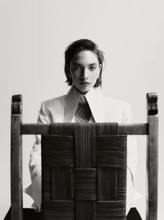 Elegant studio portrait of a stylish woman in a white blazer sitting behind a woven chair, showcasing fashion and design elements.