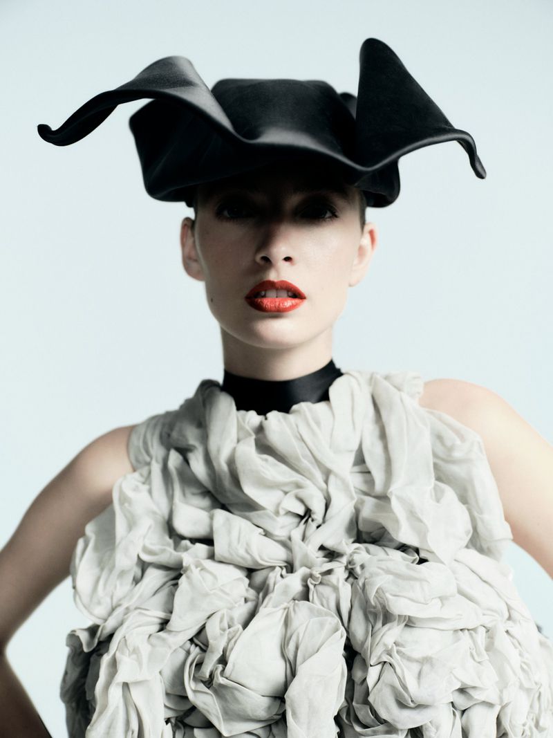 Fashion portrait of a woman with red lips wearing a draped dress and a black hat in a studio setting against a pale blue background.