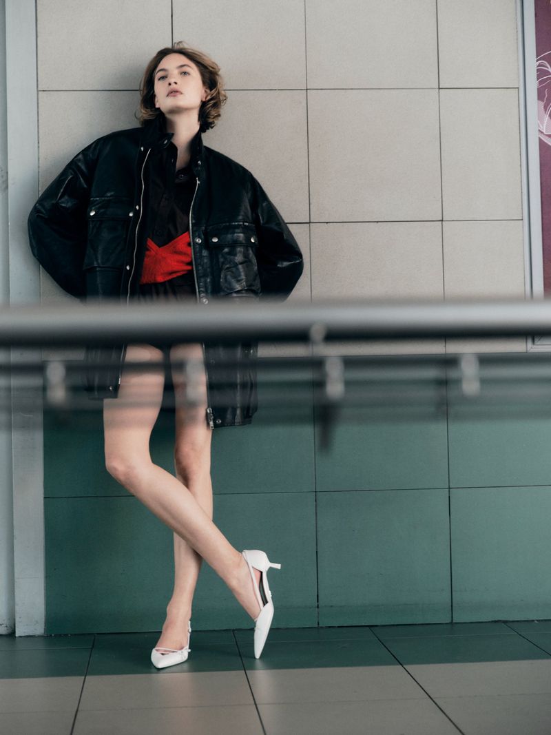 A woman in a black jacket and short dress with a red sash poses against a tiled wall, wearing white heels in a fashion forward portrait.