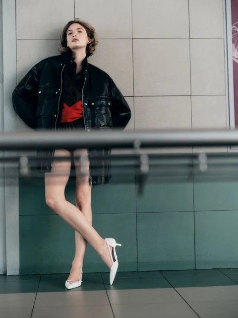 A woman in a black jacket and short dress with a red sash poses against a tiled wall, wearing white heels in a fashion forward portrait.