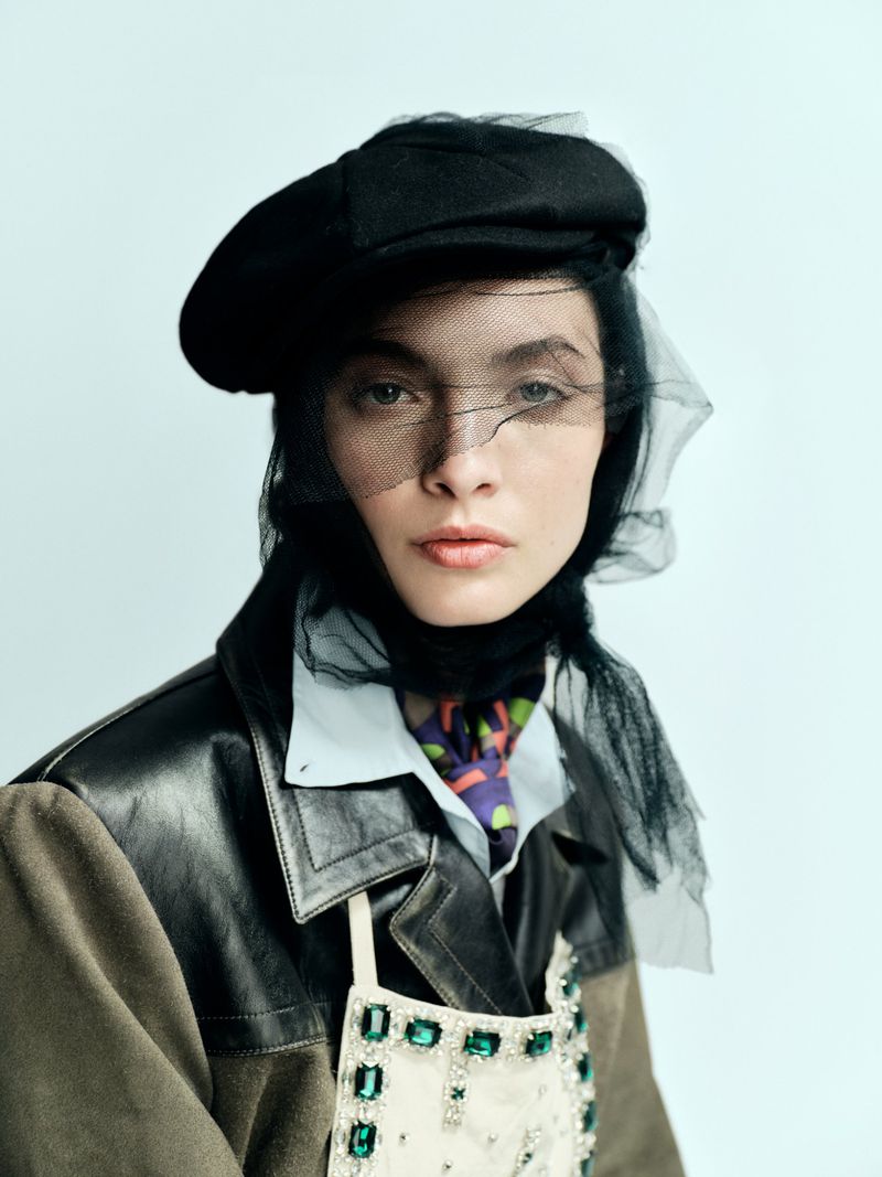 A woman in a black beret with a veil, a jacket with a cream-colored bib and jewel details poses against a light grey background.