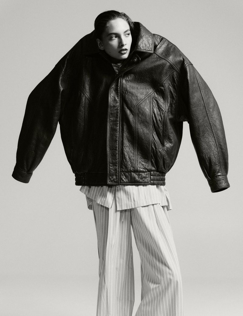 Editorial fashion shot of a female model wearing an oversized black leather jacket and striped pants against a neutral background.