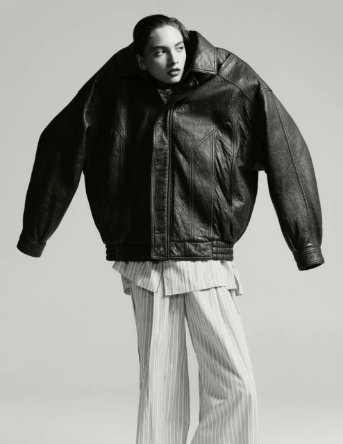 Editorial fashion shot of a female model wearing an oversized black leather jacket and striped pants against a neutral background.