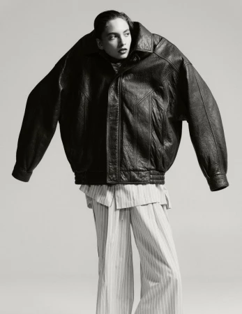 Editorial fashion shot of a female model wearing an oversized black leather jacket and striped pants against a neutral background.