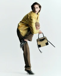 Model Rolf wearing yellow shirt and brown textured trousers holding handbag against a seamless white background