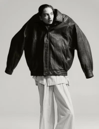 Model wearing an oversized leather jacket pulled up over the head with pinstripe trousers against a grey backdrop