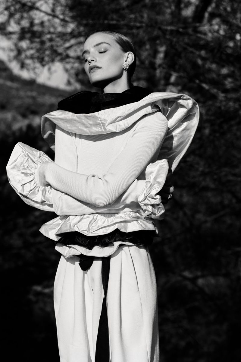 Black and white portrait of a blonde female model with closed eyes posing outdoors in a high-fashion white ruffled dress.