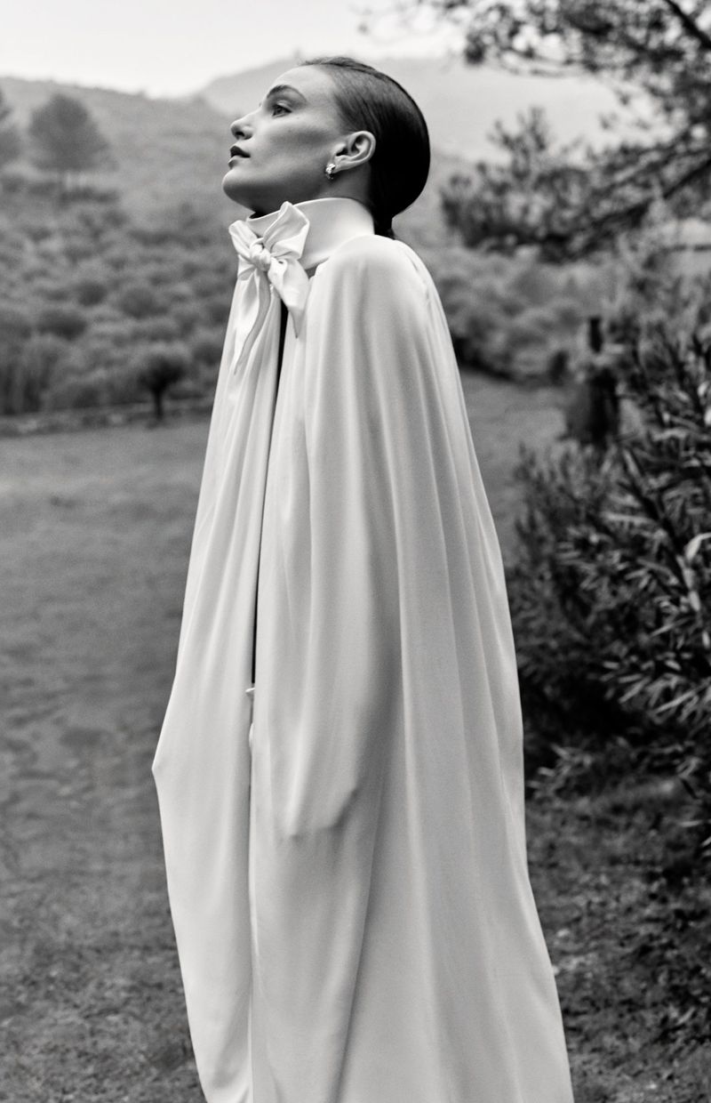 A woman in a long white gown poses in a black and white outdoor fashion shot with trees and vegetation in the background.