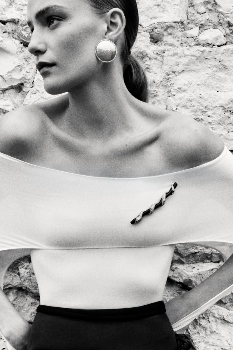 Monochrome portrait of woman in off-the-shoulder top and large disc earrings posing against a rough stone wall outdoors.