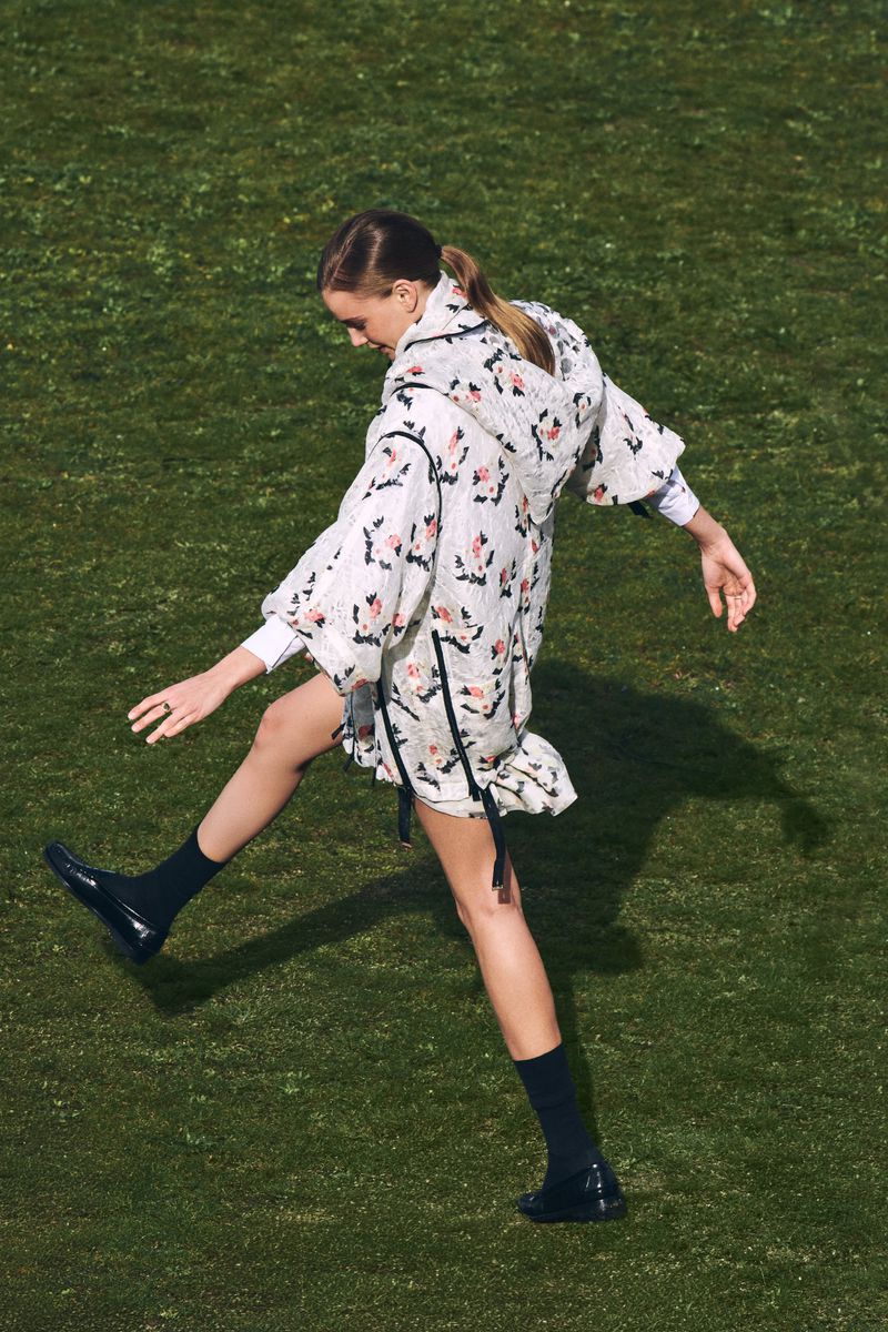 Woman in patterned coat kicks leg up on green grass outdoors. Fashion shot of adult female wearing dark socks and loafers in daytime.