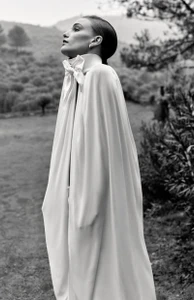 Model in a high-collared white cape dress standing outdoors in a landscape for Vogue Espana by David Portela