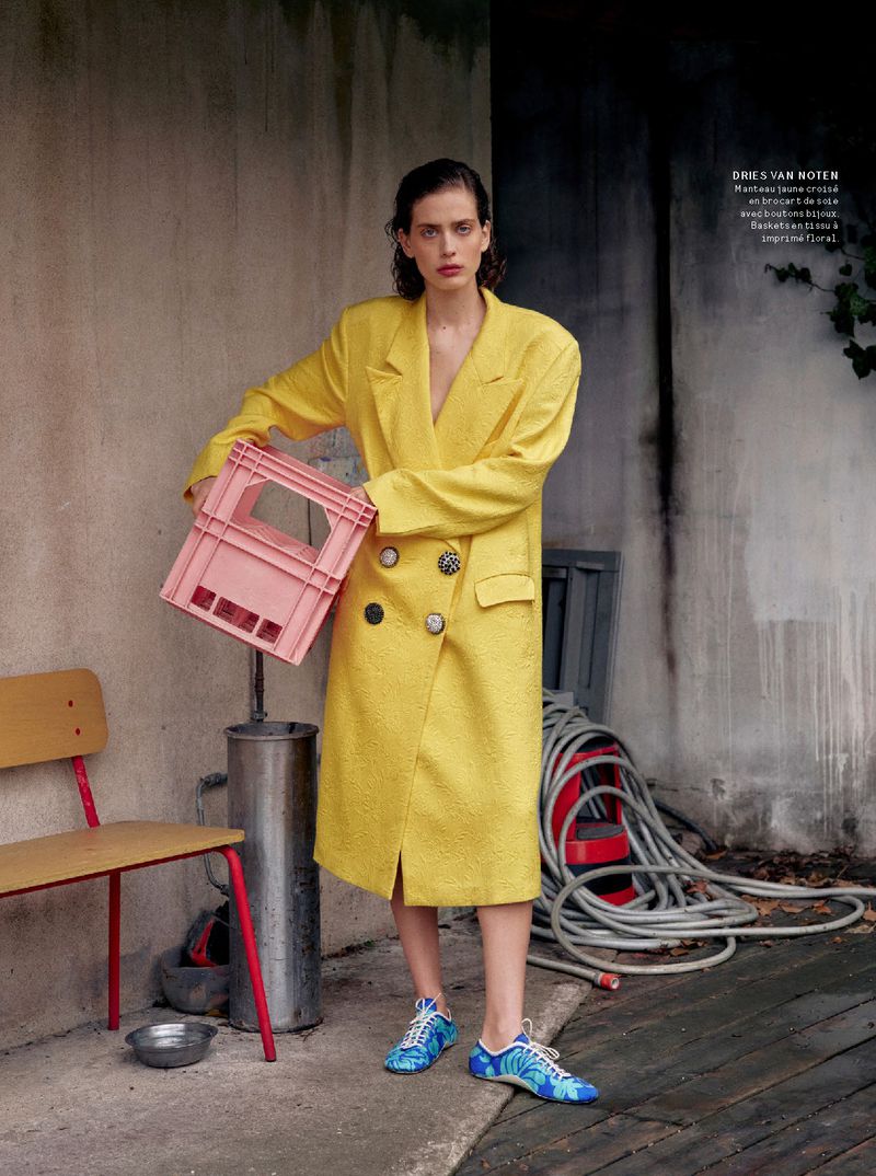 Adult female model poses outdoors wearing a yellow jacket and blue shoes, holding a pink crate, with a chair and hose in view.