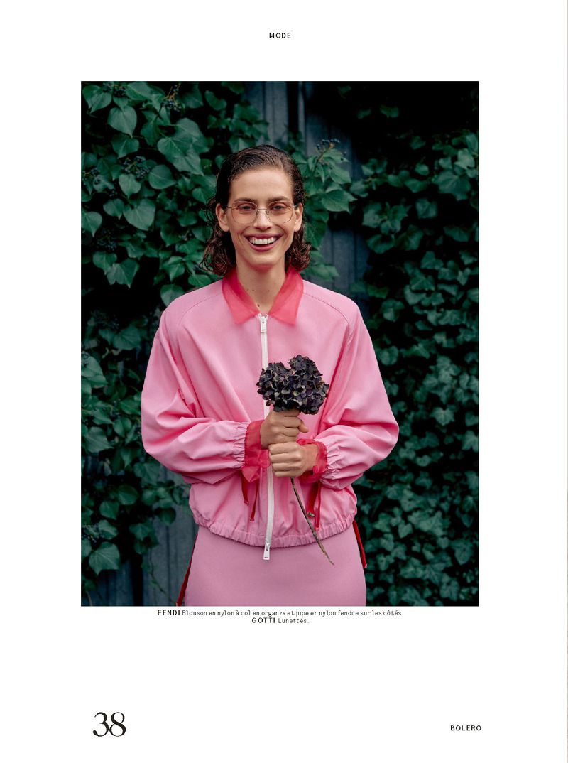 A woman with glasses smiles while holding dark flowers, wearing a pink jacket and skirt, standing against a leafy green background.