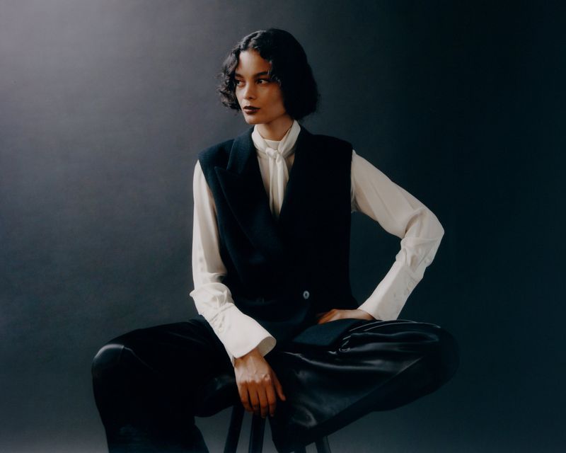 Elegant woman in a white blouse and black vest poses seated against a gray backdrop, showcasing a blend of formal and fashion-forward style.