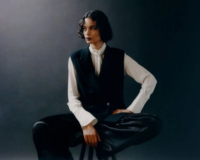 Elegant woman in a white blouse and black vest poses seated against a gray backdrop, showcasing a blend of formal and fashion-forward style.