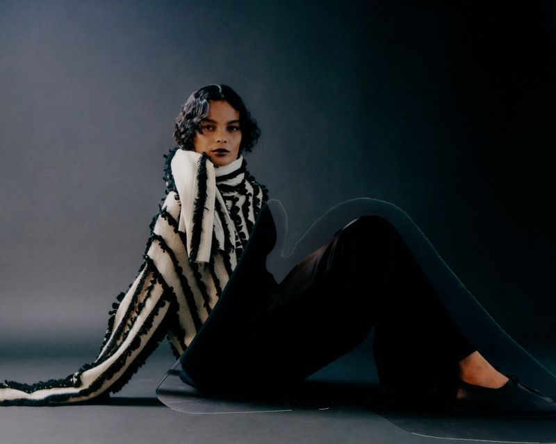 Model with dark wavy hair wearing a black and white fringed sweater sitting against a dark gradient studio background.
