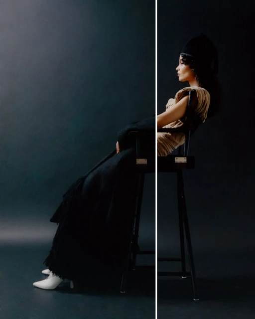 A woman sits on a chair wearing an elaborate headpiece and long black coat in a dramatically lit studio portrait.