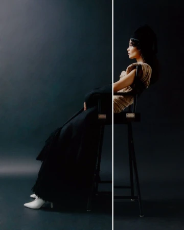 A woman sits on a chair wearing an elaborate headpiece and long black coat in a dramatically lit studio portrait.