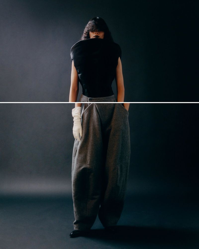 Fashionable adult poses in a sleeveless black top and grey pleated pants in a studio setting. Gloves and dark lipstick complete the style.