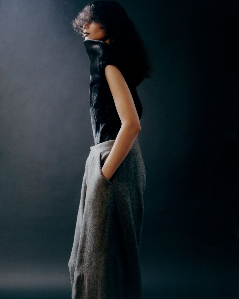 An adult female poses in profile, wearing a black top and grey trousers in a fashion studio, against a dark background with soft lighting.