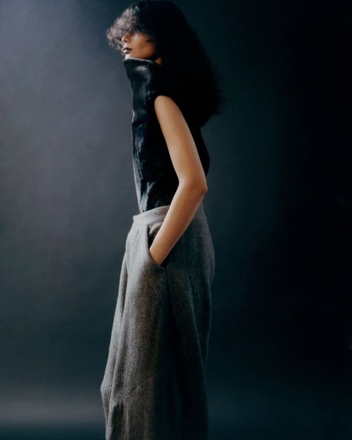 An adult female poses in profile, wearing a black top and grey trousers in a fashion studio, against a dark background with soft lighting.