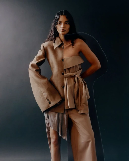 Fashion editorial shot featuring an adult female model with long curly hair posing in beige and tan clothing against a dark backdrop.