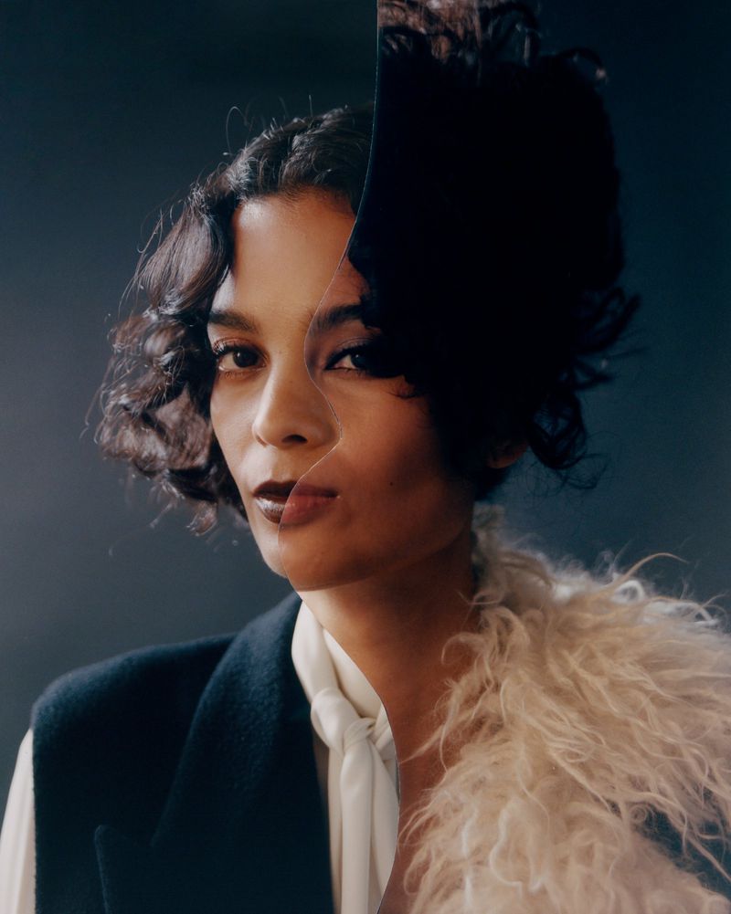 Portrait of a woman with dark curly hair wearing a white blouse and black vest against a dark background