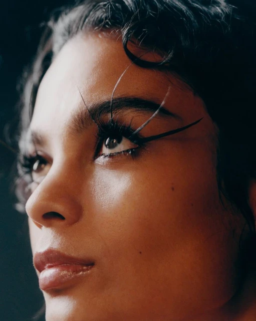 Close-up portrait of an adult female with curly hair and dramatic eye makeup, looking up against a dark backdrop in soft lighting.