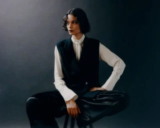 Model seated on a stool wearing a black vest, white blouse, and dark trousers against a grey gradient background.
